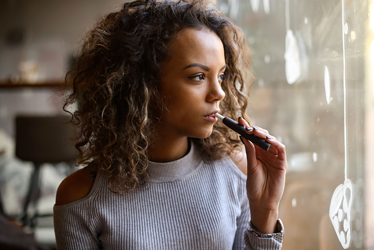 A curly-haried, dark-skinned young woman looks out a window while holding an e-cigarette to her lips. 
